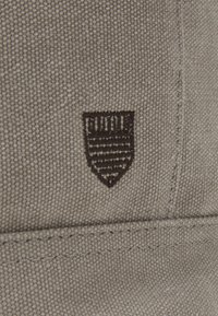 Teksturowana beżowa tkanina z haftowanym ciemno-brązowym logo tarczy, które ma poziome paski. Starannie przeszyte szwy.