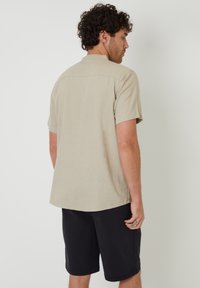 Camisa de manga corta en tono beige claro con corte recto y yugo en la espalda, combinada con pantalones cortos oscuros, de diseño sencillo y textura suave.