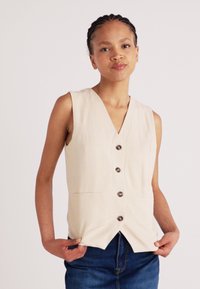 Beige ärmellose Weste mit V-Ausschnitt, fünf braunen Knöpfen, zwei seitlichen Taschen und einem leicht taillierten Saum. Über blauen Jeans getragen.