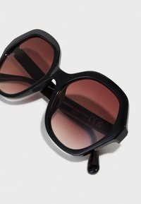 Schwarze übergroße Sonnenbrille mit braunen Verlaufslinsen, eckigem Rahmen-Design und glänzender Oberfläche. Der Text am inneren Bügel gibt Herstellungsdetails an.