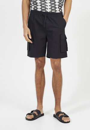Shorts cargo noirs avec poches latérales, taille élastique et cordon de serrage. Associés à des sandales noires, présentant une large lanière et un détail de boucle.