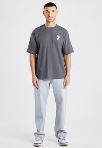 Grijs T-shirt met korte mouwen en een subtiel logo op de borst, gecombineerd met lichtblauwe, loszittende jeans en grijze sneakers.