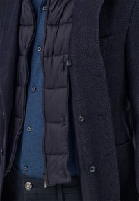 Tmavomodré štruktúrované sako s viditeľne vypchávkou vo vnútri; vrstvené cez modrú košeľu na gombíky; zapínané vpredu s viditeľnými detailmi na vreckách.