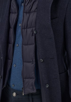 Marineblauer Blazer mit strukturierter Oberfläche und sichtbarem, gepolstertem Innenfutter; darüber getragen ein blaues Hemd mit Knöpfen; vorne zugeknöpft mit sichtbaren Taschenakzenten.