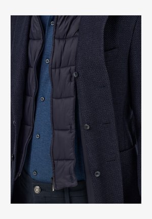 Marineblauer Blazer mit strukturierter Oberfläche und sichtbarem, gepolstertem Innenfutter; darüber getragen ein blaues Hemd mit Knöpfen; vorne zugeknöpft mit sichtbaren Taschenakzenten.