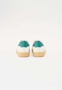 White sneakers with a green heel tab and tan rubber sole. Smooth leather material, minimalist design, and rear branding detail.
