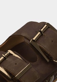 Καφέ δερμάτινο λουράκι σανδαλιού με δύο λαμπερές ορθογώνιες αγκράφες σε χρυσό τόνο και ορατές τρύπες ρύθμισης.