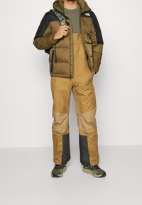 Vadderad huva jacka i brun och svart, tillsammans med bruna hängselbyxor med färgblockering och mörkgrå detaljer. Gröna sneakers.