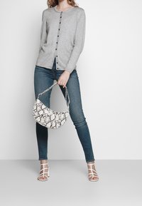 Grå knapp-up cardigan, blå jeans, vita sandaletter med remmar och en vit handväska med ormtryck i en böjd form och strukturerad yta.