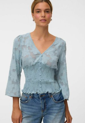 Femme portant un chemisier bleu clair transparent texturé avec des boutons et une taille smockée, associé à un jean bleu, debout devant un fond blanc.