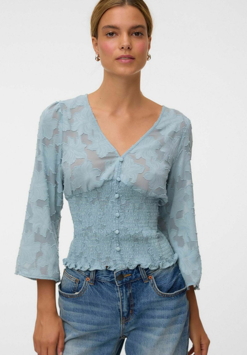 Femme portant un chemisier bleu clair transparent texturé avec des boutons et une taille smockée, associé à un jean bleu, debout devant un fond blanc.