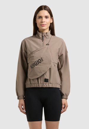 Woman wearing a beige zip-up jacket and crossbody bag with "KHUJO" text, paired with black cycling shorts, standing against a plain background.