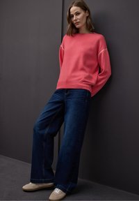 Femme appuyée contre un mur sombre portant un pull corail, un jean bleu foncé à larges jambes et des baskets beiges, regardant la caméra.