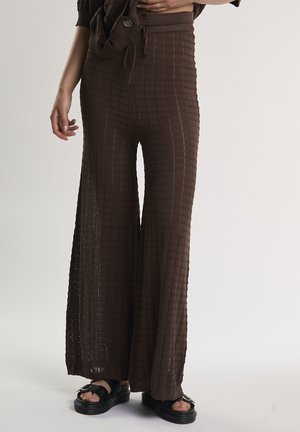 Ruskeat teksturoidut leveälahkeiset housut, joita käytetään mustien korokepohjasandaalien kanssa, vyötäröstä varpaisiin tasaisella taustalla.