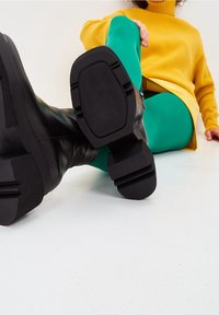 Stivaletti neri alla caviglia con finitura liscia, suole spesse e motivi testurizzati sul fondo, abbinati a leggings verdi e un maglione giallo.