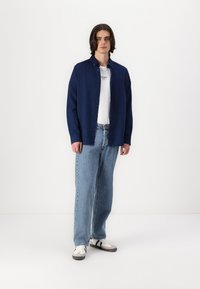 Cămașă albastră cu nasturi, dintr-un material texturat, purtată peste un tricou alb și asortată cu blugi albaștri deschis și teniși albi cu accente negre.