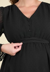 Čierne šaty so štruktúrovanou tkaninou s vyvýšenými bodkami, výstrihom do V, piatimi prednými gombíkmi a viazaním v páse. Priesvitné rukávy s čipkovými detailmi.