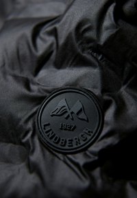 Tessuto nero con una superficie testurizzata presenta un emblema rotondo in rilievo di colore nero. L'emblema mostra bandiere incrociate e il nome del marchio "Lindbergh."
