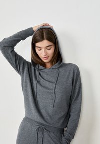 Junge Frau in einem grauen Kapuzensweater und passenden Hosen, mit einer Hand auf ihrem Kopf, die vor einer weißen Wand steht.
