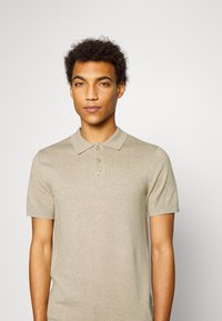 Beige kortärmad polo med ribbad textur, treknappslå och broderad logotyp på bröstet.