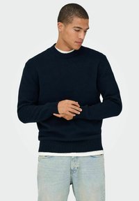 Maglione a maglia blu navy con scollo rotondo, polsini e bordo a coste; morbido al tatto, maniche lunghe, indossato sopra una camicia bianca.