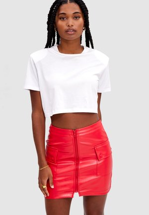 Femme aux cheveux tressés portant un t-shirt blanc court et une mini-jupe en fausse cuir rouge avec fermeture éclair à l'avant et poches à rabat.