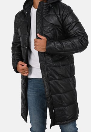 Mann trägt einen langen schwarzen gesteppter Mantel mit Reißverschluss und Druckknöpfen über einem weißen Hemd und blauen Jeans und richtet die Vorderseite des Mantels.