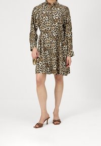 Donna che indossa un mini abito a maniche lunghe con stampa leopardata, sandali con tacco marroni e borchie, mentre tiene una piccola borsa dorata.