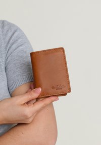 Brązowy skórzany portfel ze stębnowanymi brzegami, składany w dwa. Gładka tekstura i kompaktowy rozmiar, trzymany między palcami.