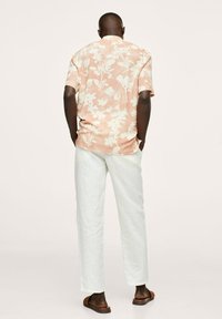 Homme portant une chemise à manches courtes à motifs floraux pêche, pantalon blanc et sandales marron, debout les mains dans les poches, de dos sur un fond neutre.