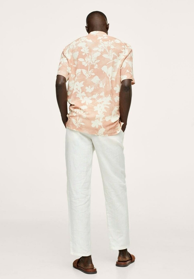 Homme portant une chemise à manches courtes à motifs floraux pêche, pantalon blanc et sandales marron, debout les mains dans les poches, de dos sur un fond neutre.