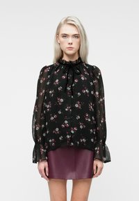 Giovane donna che indossa una blusa nera trasparente con motivo floreale, colletto e polsini a volant, abbinata a una minigonna in pelle di un viola scuro, in piedi.