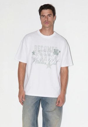 Jeune homme portant un T-shirt blanc oversize avec un texte gris clair et un jean bleu délavé, debout devant un fond blanc uni.