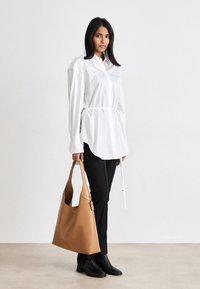 Leren handtas in cognackleur met een nonchalant ontwerp en een enkele riem. Model draagt een wit overhemd met knopen en een gerende taille, gecombineerd met zwarte broek.