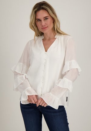 Vrouw met blond haar die een wit blouseje met knopen en ruches aan de mouwen draagt en donkerblauwe jeans, staand tegen een neutrale achtergrond.