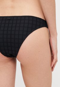 Black bikini bottom with textured pattern, featuring a low-rise cut and adjustable elastic waistband. Material appears soft and flexible.