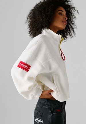 Biely flísový pulóver s vysokým golierom, žltým zipsom, červeným sťahovacím šnúrkou a červenou značkou na rukáve; v kombinácii s tmavými džínsami.