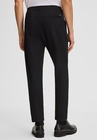 Mężczyzna w czarnych, fakturowanych spodniach, czarnych eleganckich butach i białej koszuli, pokazany od tyłu na jednolitym tle.