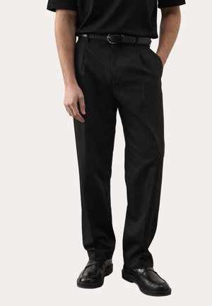 Mężczyzna ubrany w czarne spodnie o prostych nogawkach, czarny pasek, czarną koszulę z krótkim rękawem i czarne eleganckie buty, stoi z jedną ręką w kieszeni.