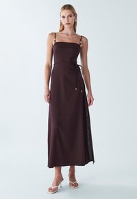 Abito lungo marrone senza maniche con scollo dritto e cintura in vita. Presenta uno spacco laterale e spalline regolabili con dettagli in perline.