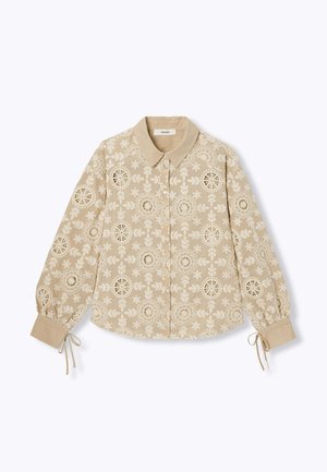 Blouse beige à manches longues avec découpes en dentelle florale, fermeture boutonnée devant, col pointu et liens aux poignets.