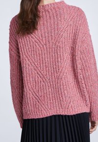 Rosa gestrickter Pullover mit einem strukturierten Muster und ribbeltem Halsausschnitt. Das Design bietet diagonale Linien und einen lockeren Schnitt, kombiniert mit einem schwarzen plissierten Rock.