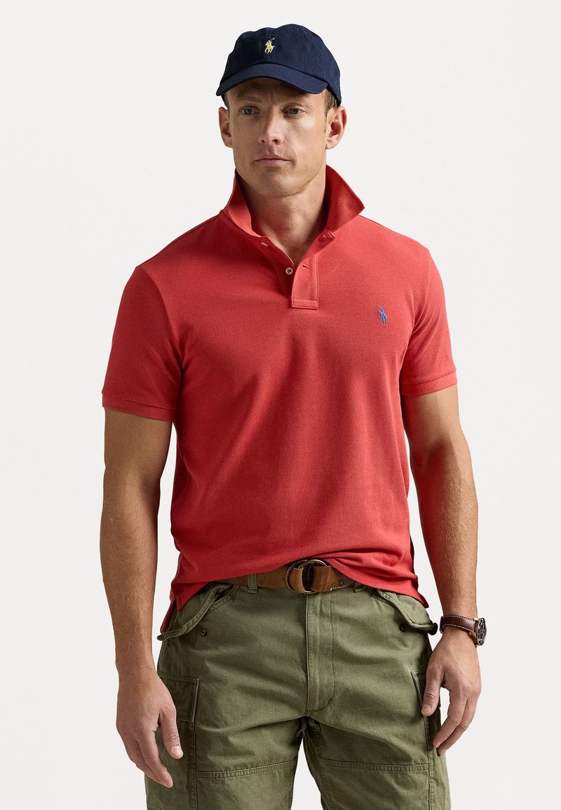 Röd polo med nedvikt krage och blå logotyp, ihop med olivgröna cargobyxor. Modellen bär en marinblå keps och ett brunt bälte.