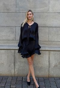 Melnās plīvētās kleitas ar garām piedurknēm, sasietu kakla izgriezumu un ruffled apakšējo malu. Apvienota ar melniem augstpapēžu kurpēm ar norādītu purngalu. Gluda, spoguļota auduma.