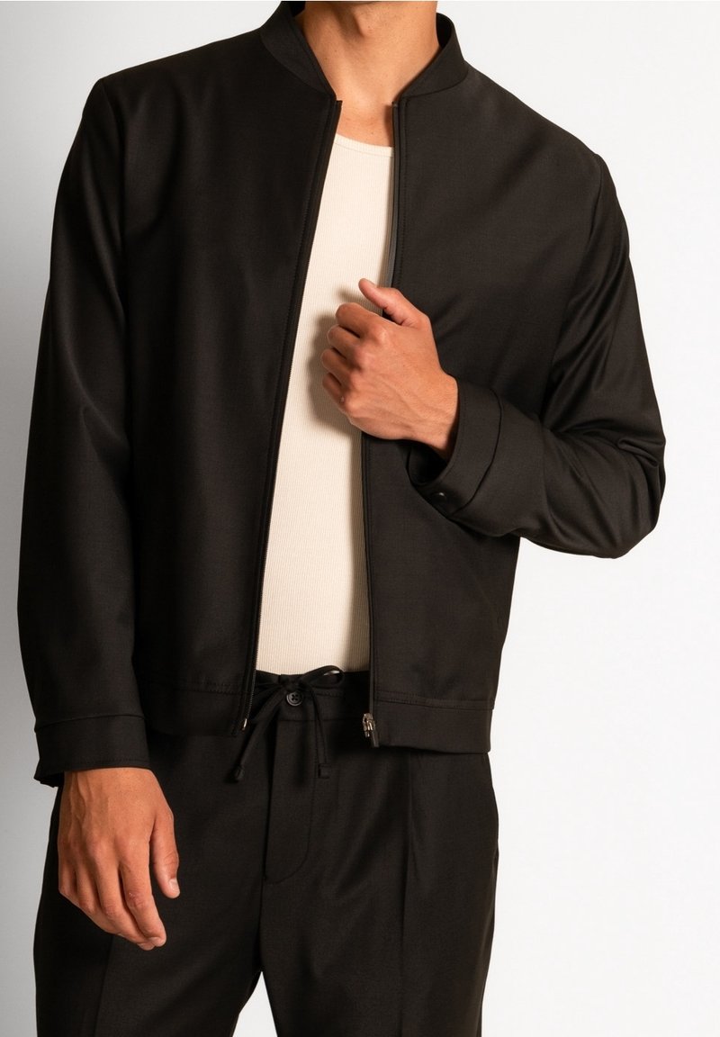 Czarna kurtka na zamek z stójką, wykonana z gładkiego materiału. Cechuje się luźnym krojem i minimalistycznym designem, noszona na jasnym topie.