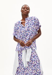 Frau trägt ein lockeres, blumiges Oberteil und einen passenden Rock in lila, rosa und blauen Tönen, mit einem weißen Hemd, das um ihre Taille gebunden ist.