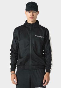 Czarna kurtka zapinana na zamek błyskawiczny z białymi paskami po bokach, z kołnierzykiem typu stojącego i elastycznymi mankietami. Materiał wydaje się gładki z delikatnym połyskiem.