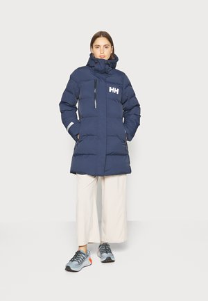 Navy puffer jacket with a high collar, zippered front, padded design, and white logo. Paired with light trousers and gray athletic shoes.