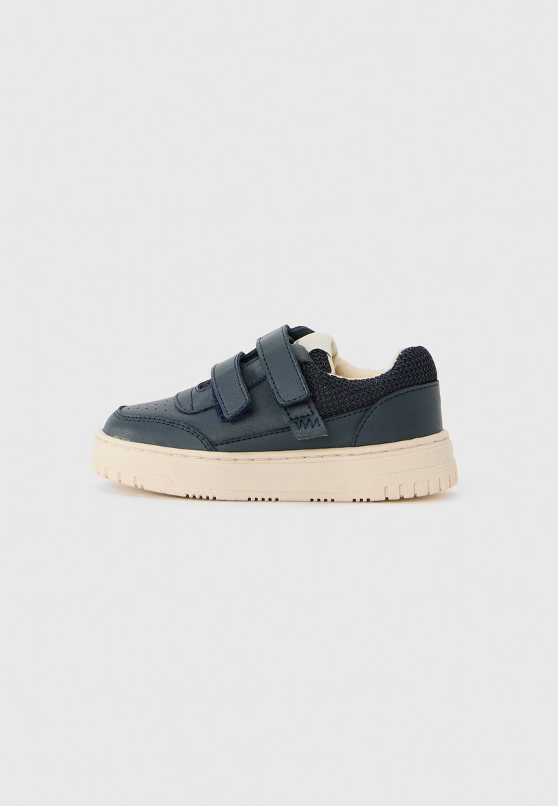 Marinblå sneaker med beige sula, två kardborreband och meshtyg vid hälen, visad från sidan på en enkel bakgrund.