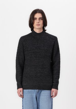 Chunky-strikket sort sweater med høj ribstrikket krave og lange ærmer, der har et struktureret mønster og ribbede manchetter. Båret med lyseblå jeans.